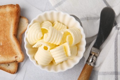 Tasty butter curls in bowl, knife and toasts on light grey table, top view