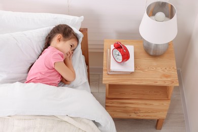 Photo of Little girl lying in bed and looking at alarm clock on bedside table indoors, above view