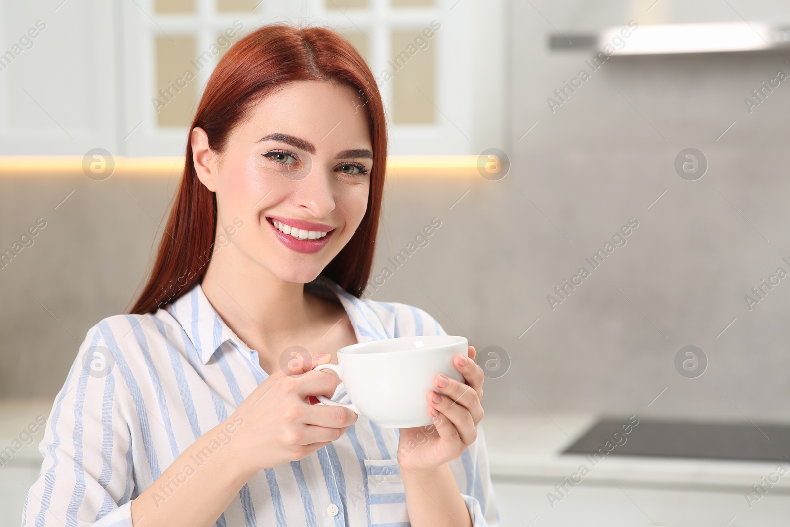 Photo of Beautiful woman with red dyed hair holding cup of drink in kitchen, space for text