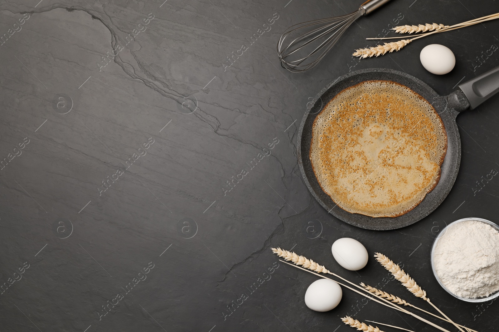 Photo of Frying pan with delicious crepe, spikelets and ingredients on black table, flat lay. Space for text