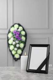 Photo of Photo frame with black ribbon, burning candle on light grey table and wreath of plastic flowers near wall indoors. Funeral attributes