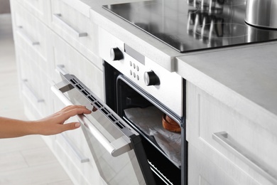 Young woman opening electric oven in kitchen, closeup