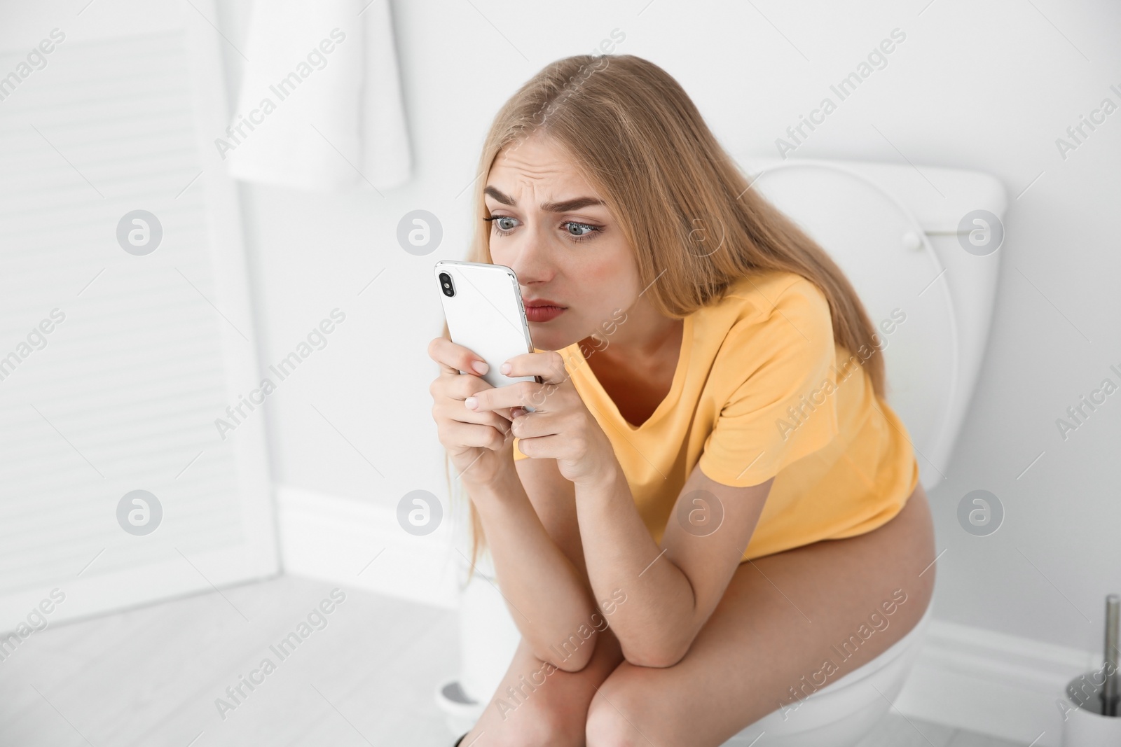 Photo of Young woman using mobile phone while sitting on toilet bowl at home