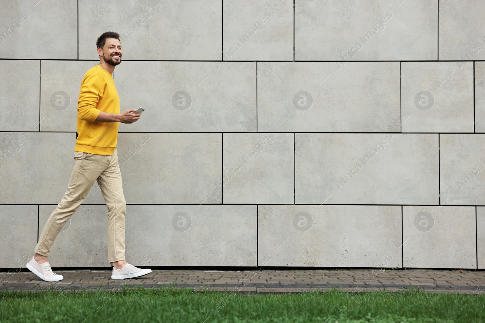 Photo of Handsome man with smartphone walking near grey stone wall outdoors, space for text