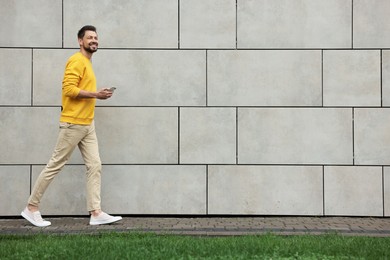 Handsome man with smartphone walking near grey stone wall outdoors, space for text