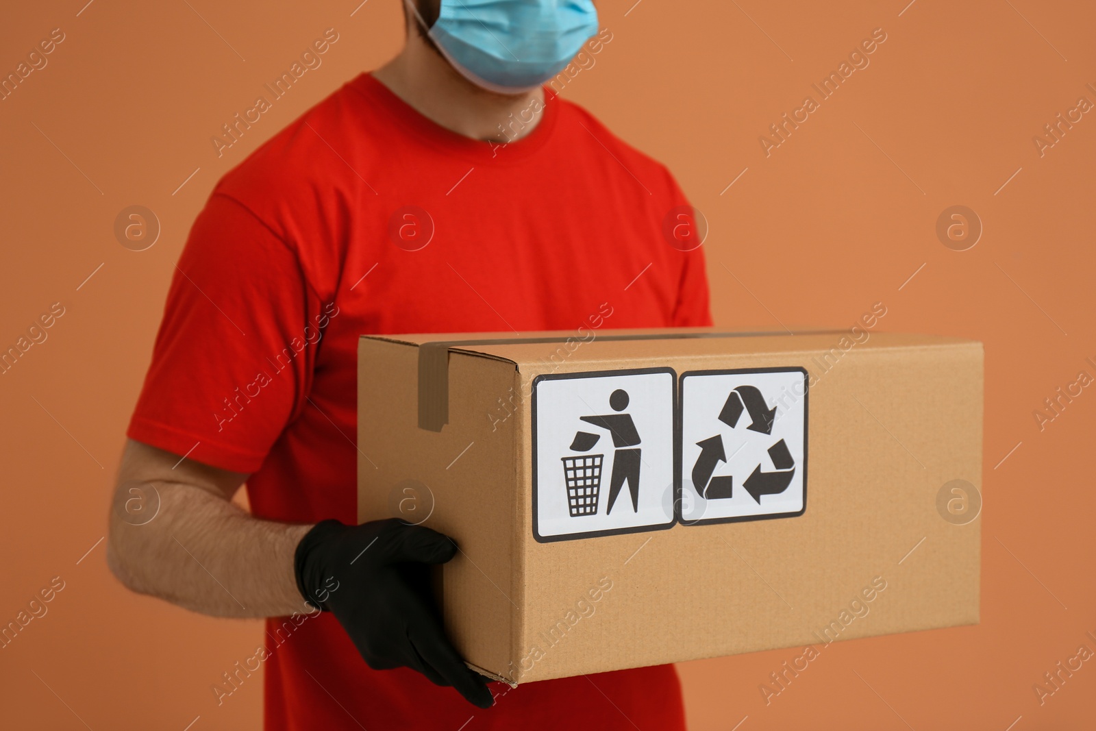 Photo of Courier holding cardboard box with different packaging symbols on orange background, closeup. Parcel delivery