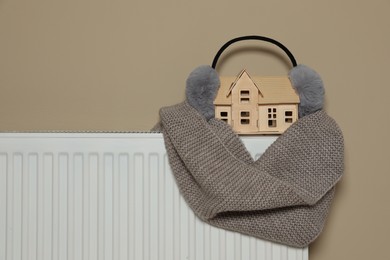 Photo of Modern radiator with earmuffs, scarf and wooden house near beige wall indoors, space for text. Winter heating efficiency