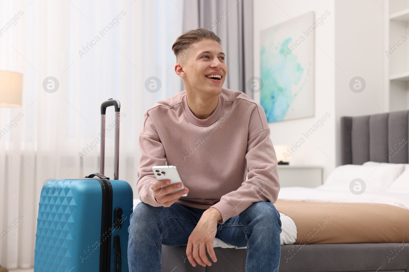 Photo of Smiling guest with smartphone relaxing on bed in stylish hotel room