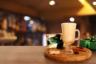 Image of Glass of delicious eggnog, spices, cookie and gift boxes on wooden table in bar, space for text