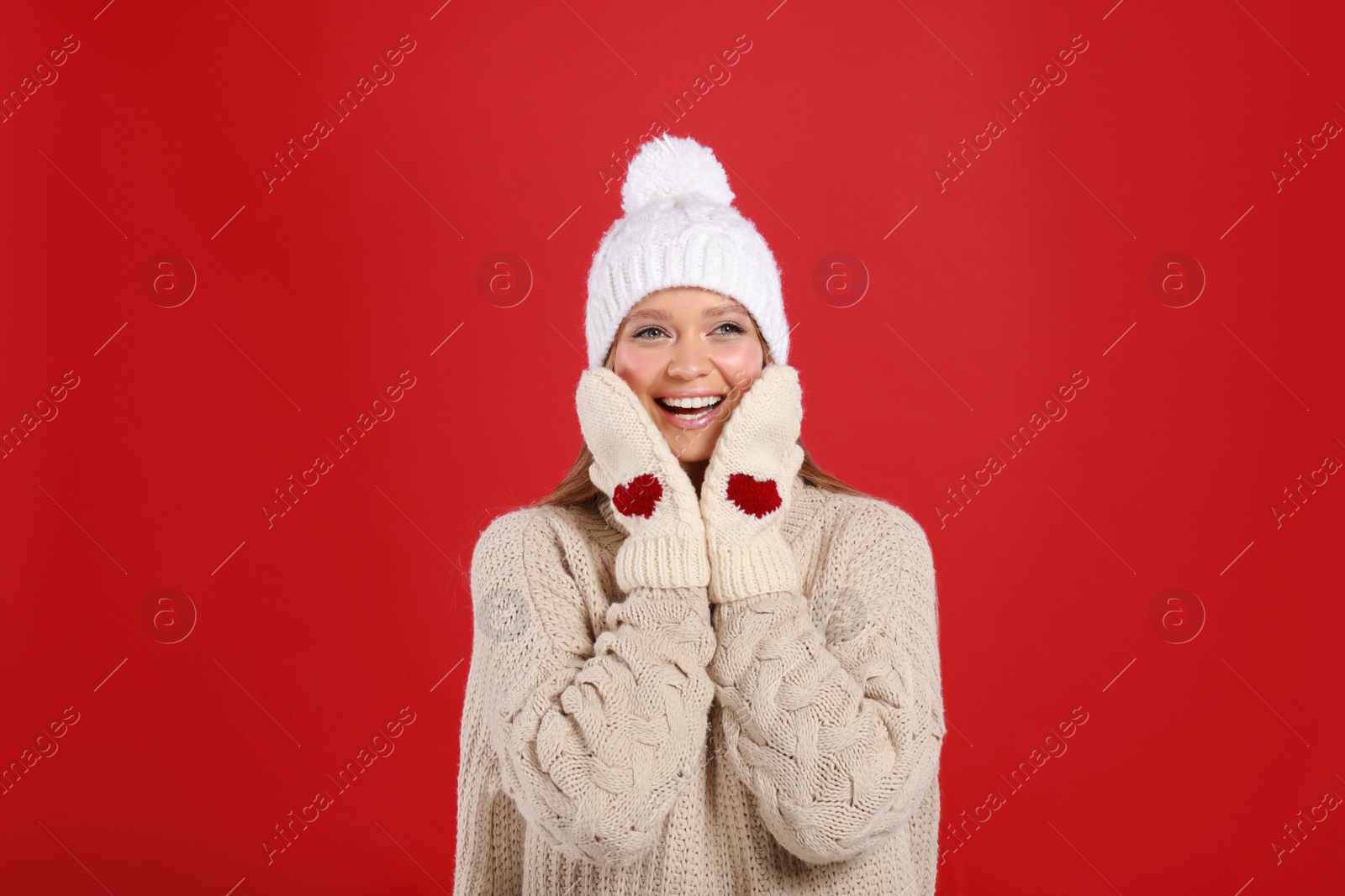 Photo of Emotional young woman in warm sweater, mittens and hat on red background. Winter season