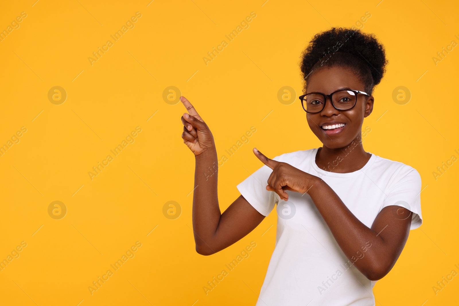 Photo of Happy young woman in eyeglasses pointing at something on orange background. Space for text