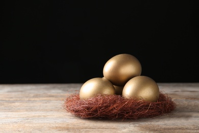Photo of Golden eggs in nest on wooden table against black background, space for text