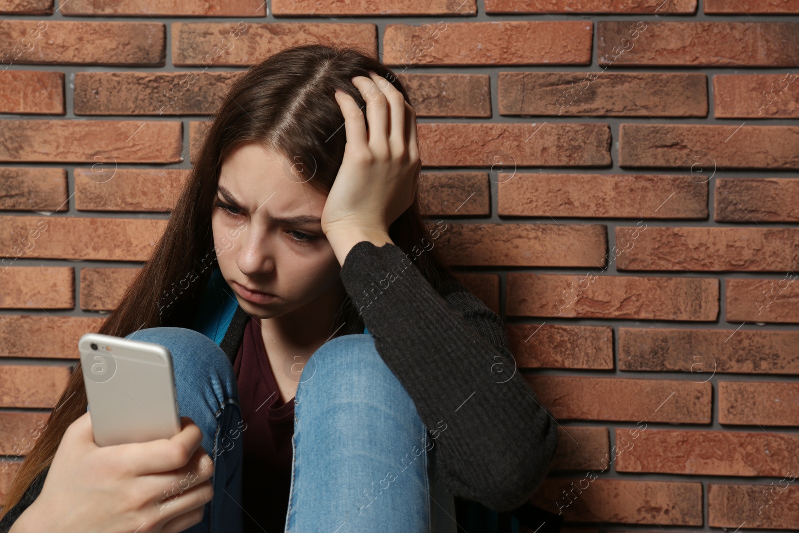 Photo of Upset teenage girl with smartphone sitting at wall indoors. Space for text