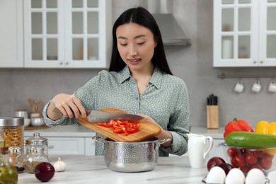 Photo of Cooking process. Beautiful woman adding cut bell pepper into pot in kitchen