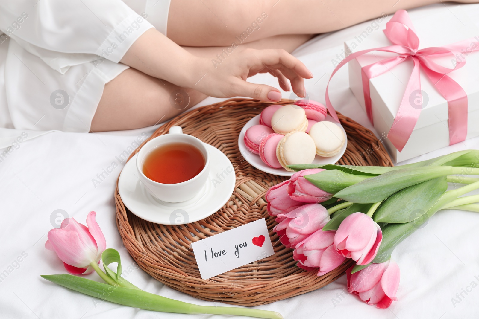 Photo of Tasty breakfast served in bed. Woman with macarons, tea, gift box, flowers and I Love You card at home, closeup