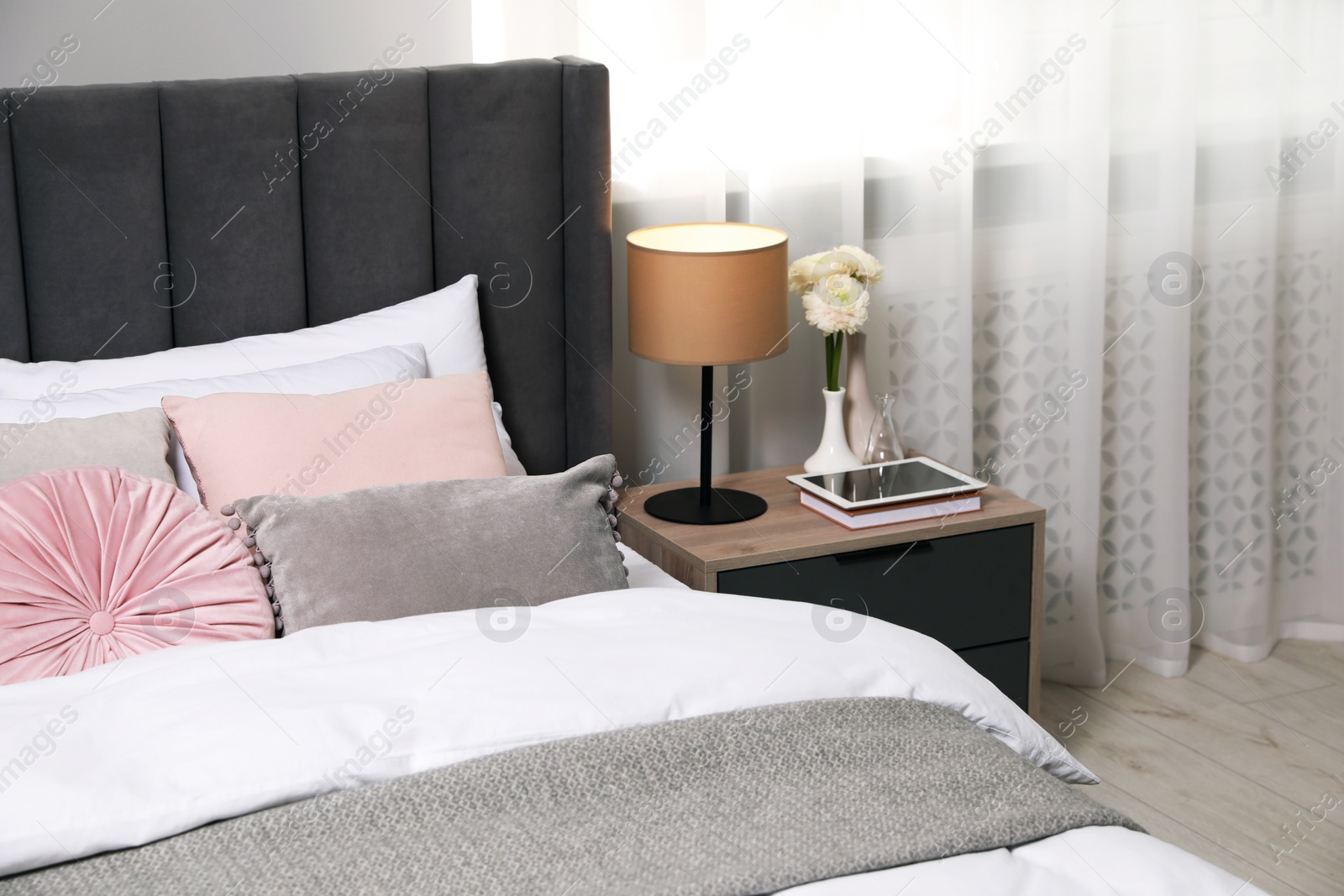 Photo of Comfortable bed with cushions, lamp and different decor on bedside table in room. Stylish interior