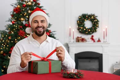 Photo of Happy young man in Santa hat opening Christmas gift at table in decorated room