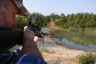 Man aiming with hunting rifle near lake outdoors, closeup. Space for text