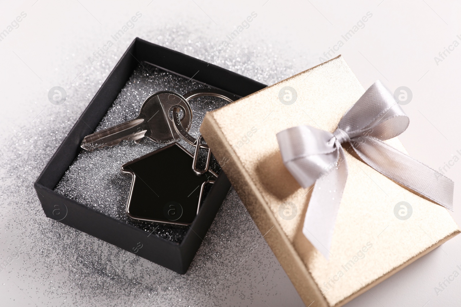 Photo of Key with trinket in shape of house, glitter and gift box on light grey background, closeup. Housewarming party
