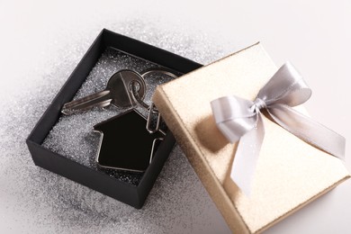 Photo of Key with trinket in shape of house, glitter and gift box on light grey background, closeup. Housewarming party