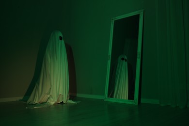 Creepy ghost. Woman covered with sheet looking at mirror in green light