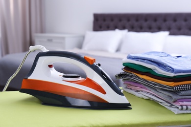 Photo of Modern electric iron and clean folded clothes on board in bedroom. Laundry day