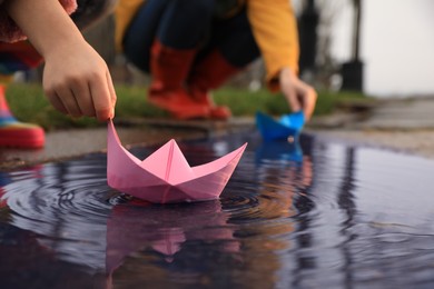 Photo of Little girl and her mother playing with paper boats near puddle outdoors, closeup
