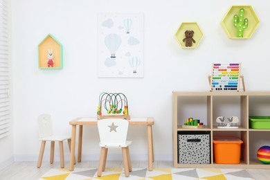 Photo of Table with chairs, shelves and toys near white wall in playroom. Stylish kindergarten interior