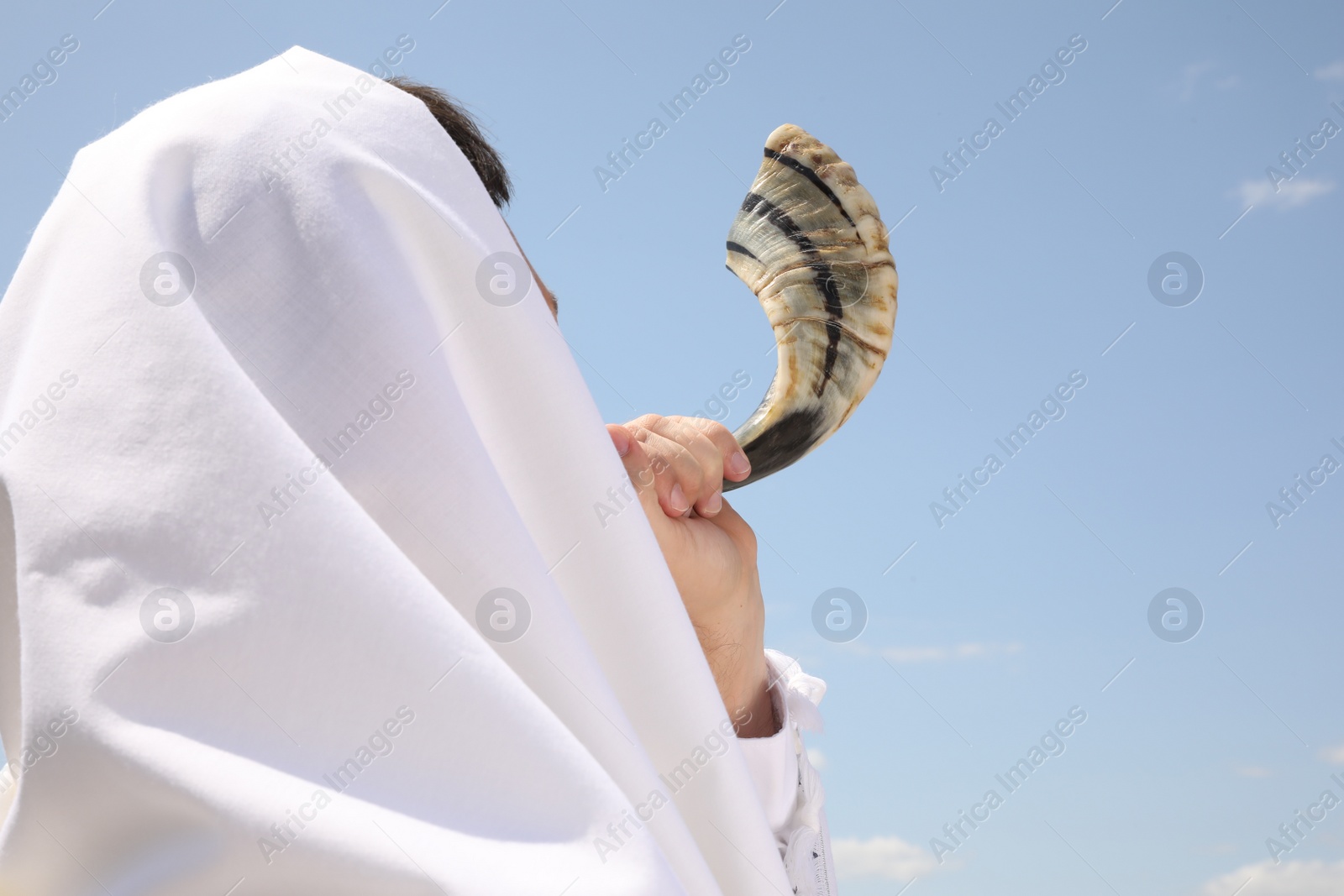 Photo of Jewish man in tallit blowing shofar outdoors. Rosh Hashanah celebration