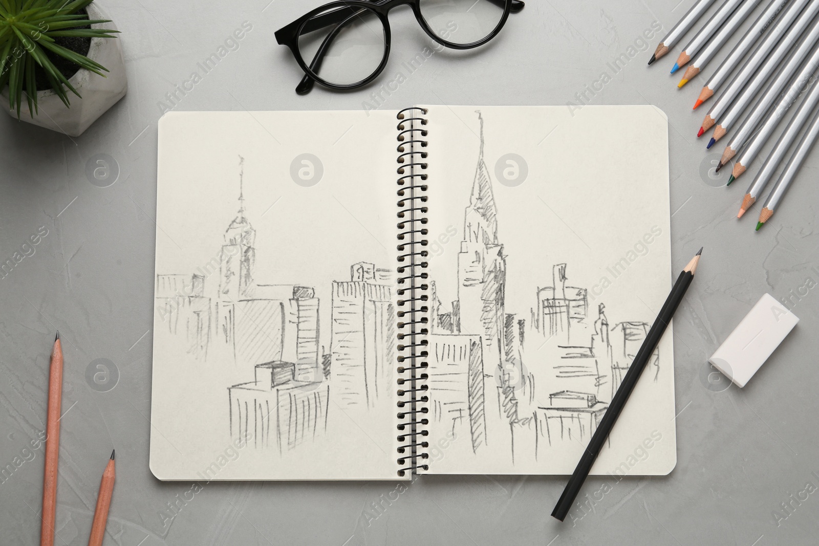 Image of Sketch of cityscape in notebook, pencils, eraser and glasses on grey table, flat lay