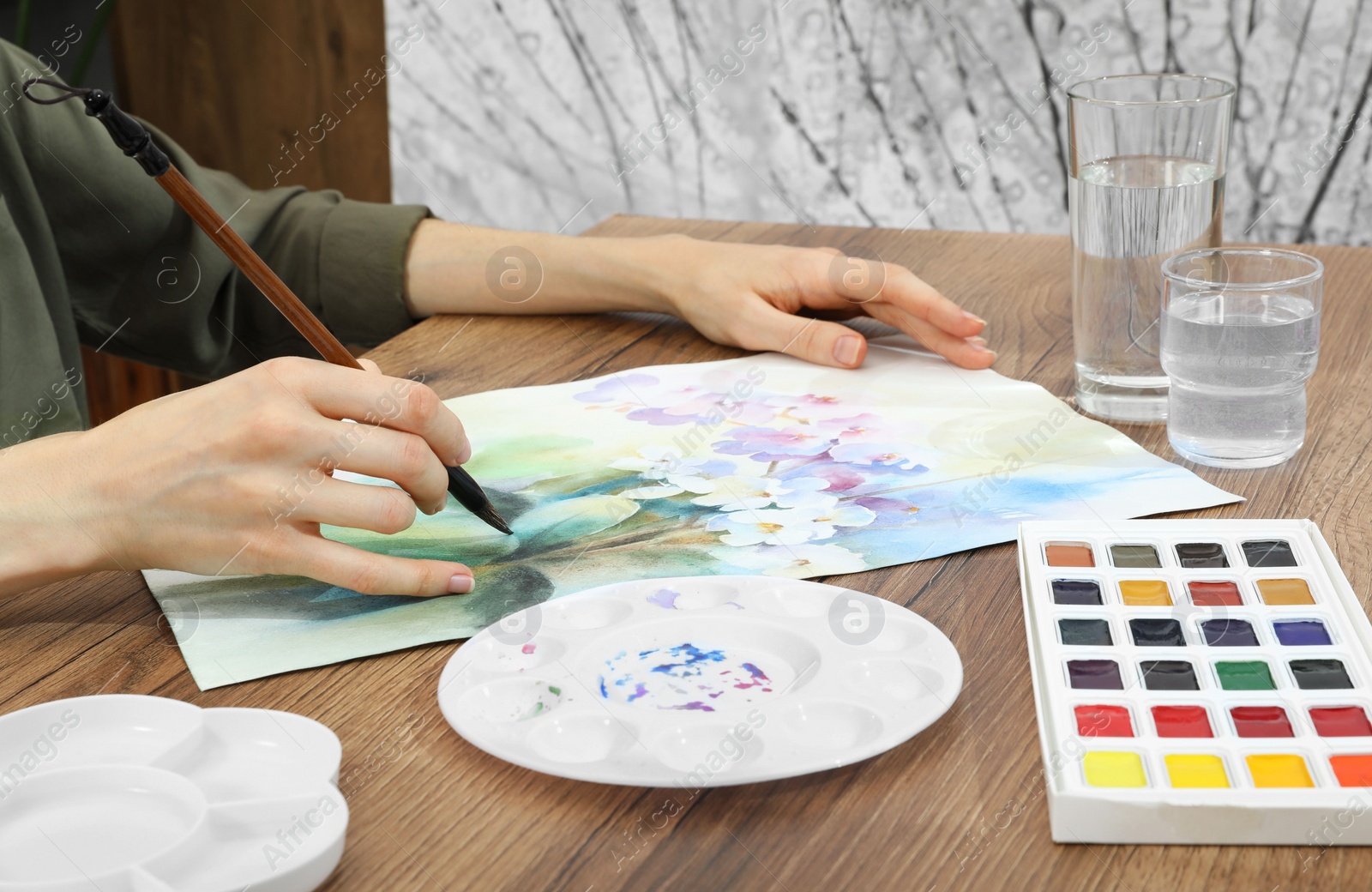 Photo of Woman painting flowers with watercolor at wooden table, closeup. Creative artwork