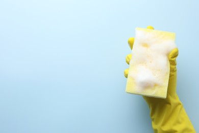 Photo of Cleaner in rubber glove holding sponge with foam on light blue background, top view. Space for text