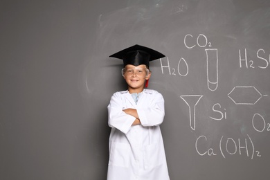 Photo of Little school child in laboratory uniform with graduate cap and chemical formulas on grey background