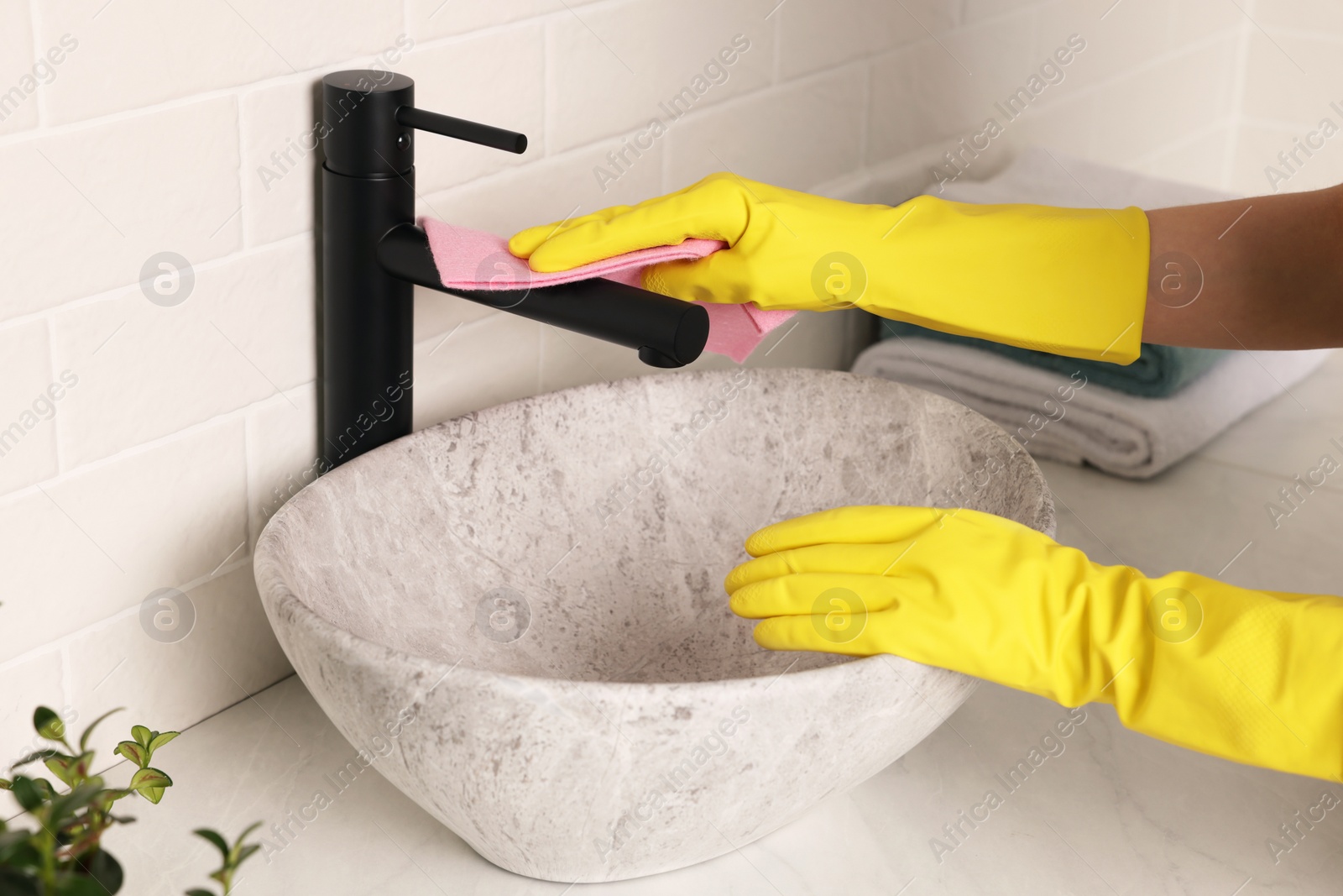 Photo of Woman in gloves cleaning faucet of bathroom sink with rag, closeup