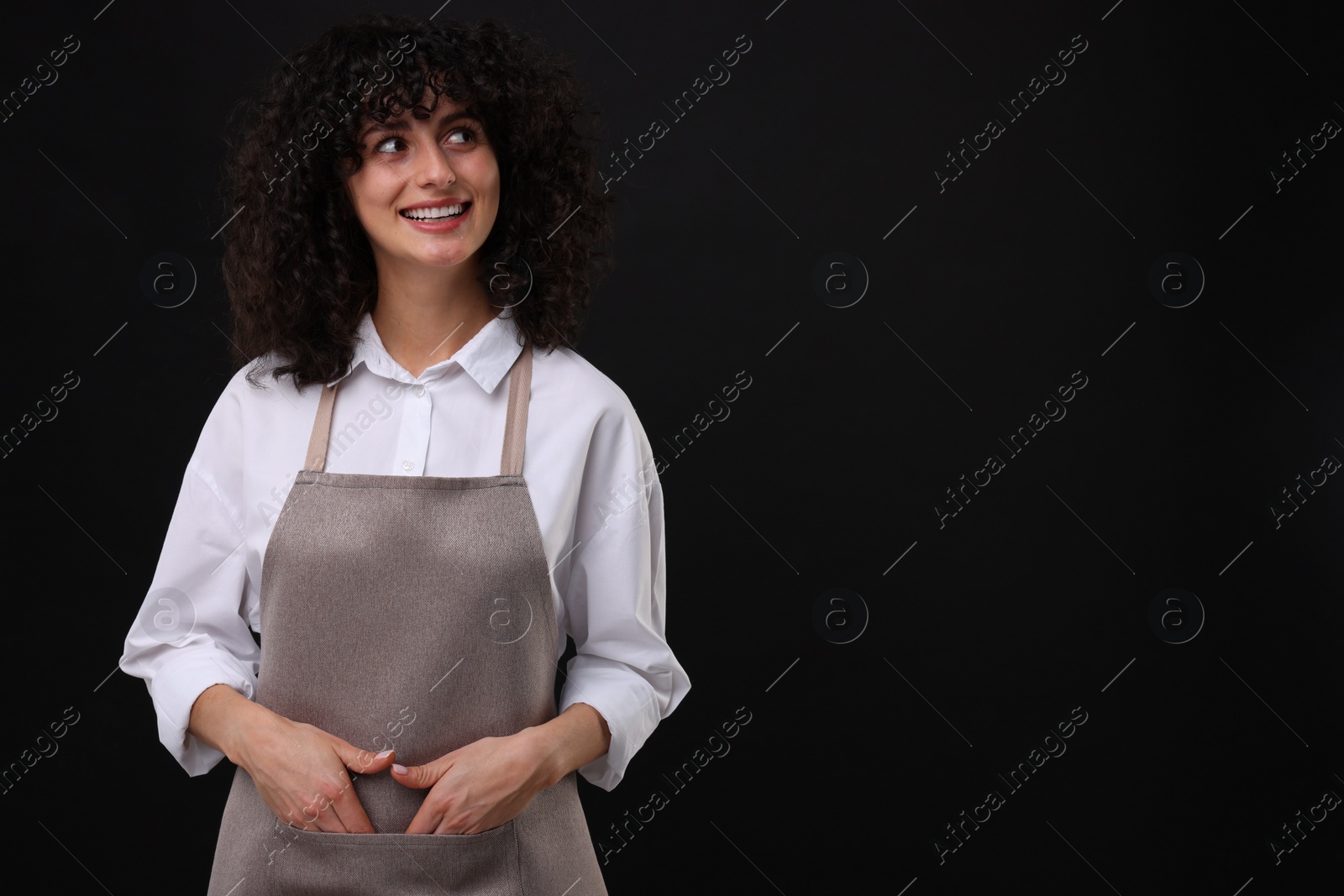 Photo of Happy woman wearing kitchen apron on black background, space for text. Mockup for design