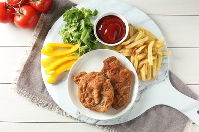Tasty schnitzels served with potato fries, ketchup and vegetables on white wooden table, flat lay