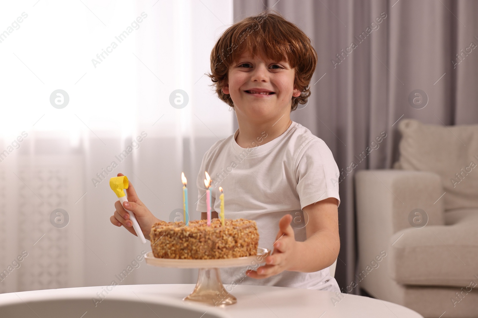 Photo of Birthday celebration. Cute boy with party blower at table with tasty cake indoors