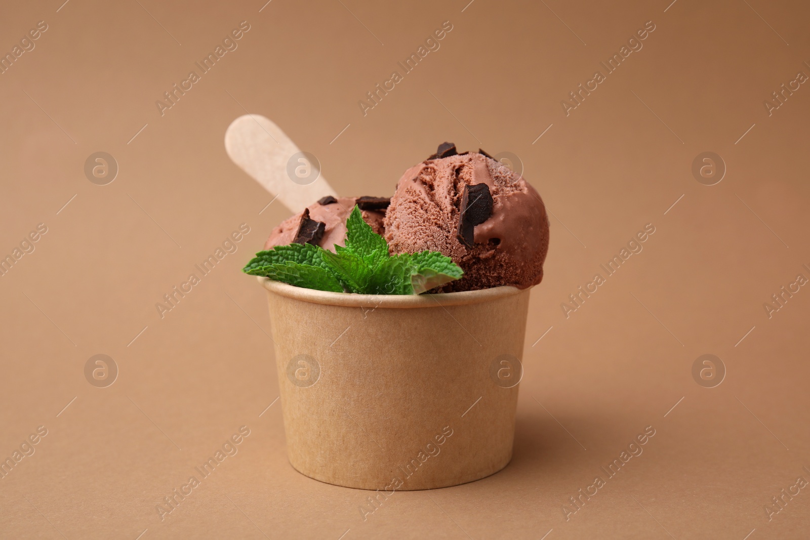 Photo of Paper cup with tasty chocolate ice cream, sticks and mint leaves on light brown background