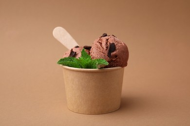 Paper cup with tasty chocolate ice cream, sticks and mint leaves on light brown background