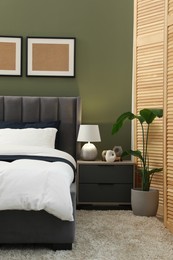 Comfortable bed, nightstand, lamp and houseplant in stylish room. Interior design