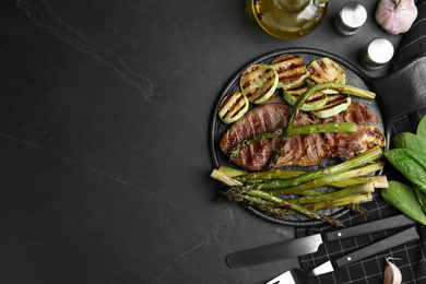 Tasty grilled meat served with asparagus and zucchini on black table, flat lay. Space for text