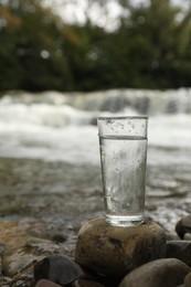 Glass of fresh water on stone near river, space for text