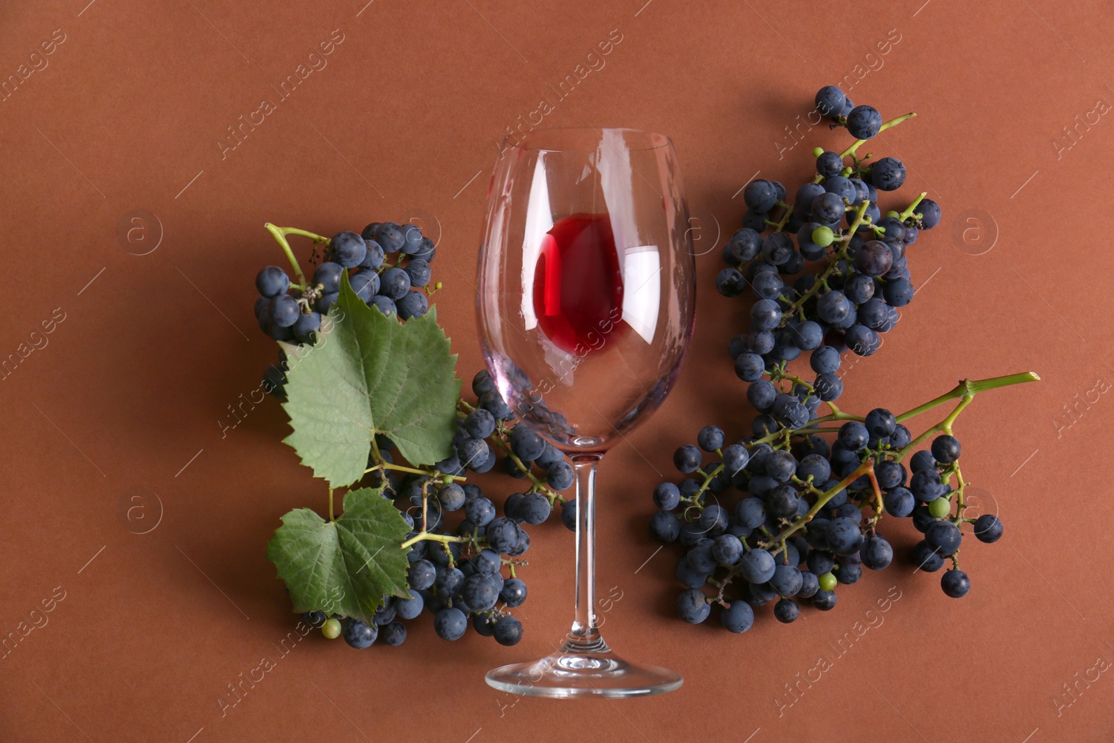 Photo of Overturned glass with red wine and grapes on brown background, flat lay