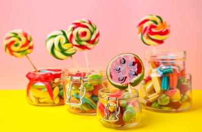 Photo of Tasty colorful candies in glass jars on yellow table against pink background