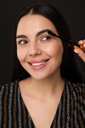 Beautiful young woman applying mascara on black background, closeup