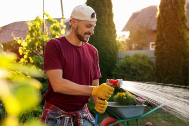Photo of Happy man watering plants from hose outdoors on sunny day. Gardening time