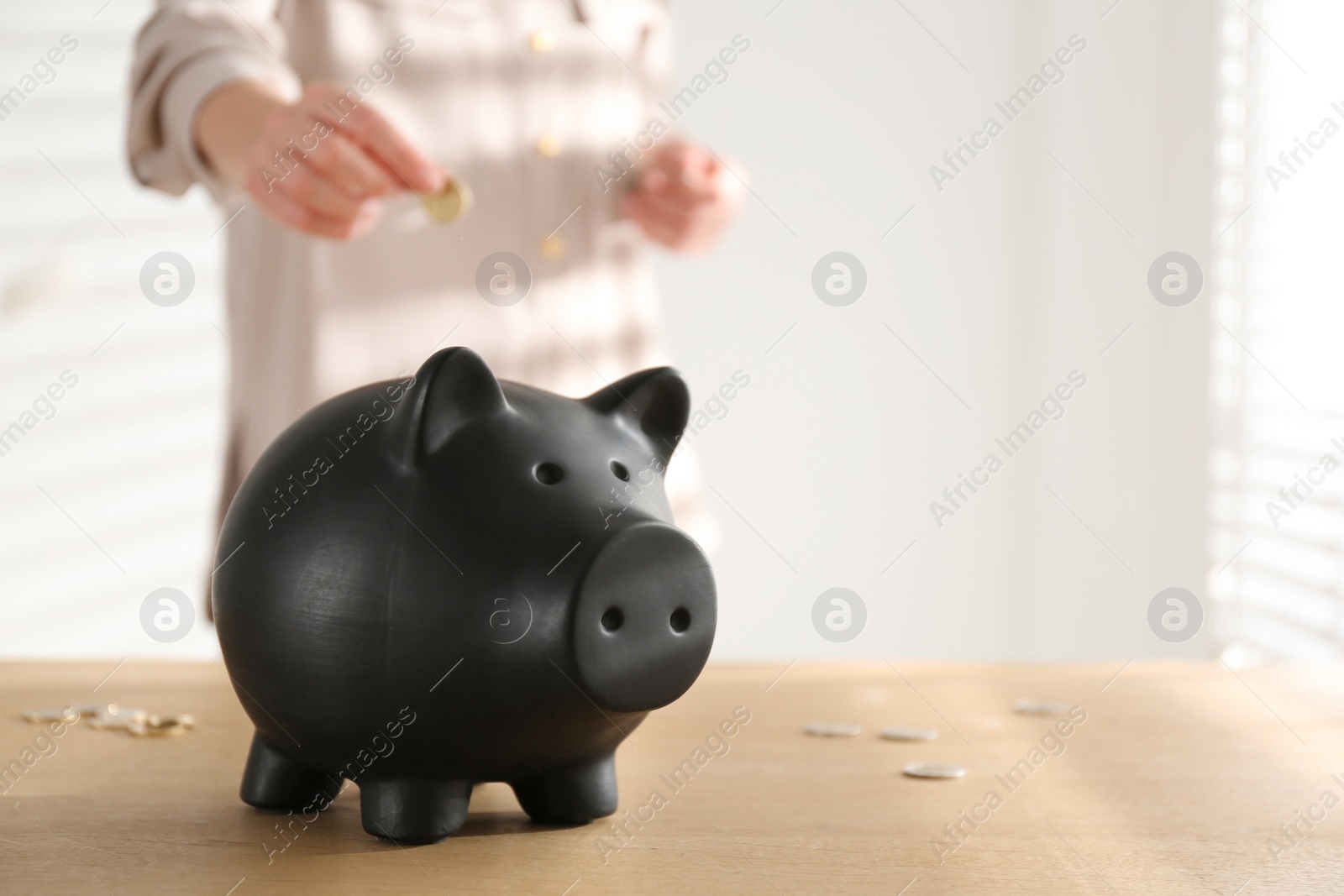 Photo of Woman putting money into piggy bank at wooden table indoors, closeup. Space for text