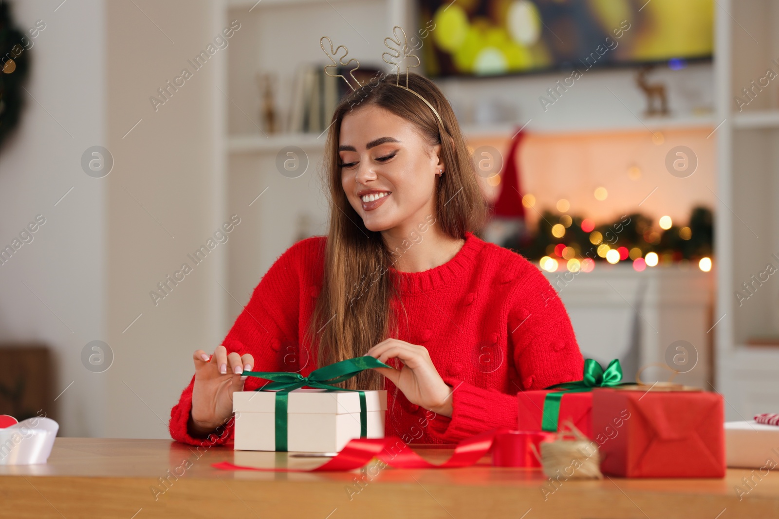 Photo of Beautiful young woman in deer headband decorating Christmas gift at table in room