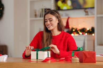 Photo of Beautiful young woman in deer headband decorating Christmas gift at table in room