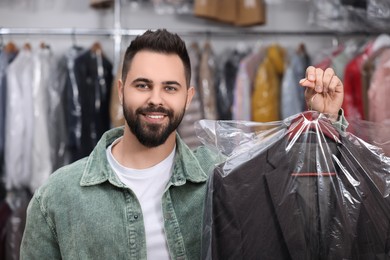 Dry-cleaning service. Happy man holding hanger with jacket in plastic bag indoors